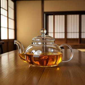Könnte beinhalten: Eine transparente Glas-Teekanne gefüllt mit bernsteinfarbenem Tee. Die Teekanne hat einen runden Körper, einen gebogenen Ausguss und einen Schlaufengriff. Der Deckel ist ebenfalls aus Glas, mit einem kleinen Knopf oben. Die Teekanne steht auf einem Holztisch.