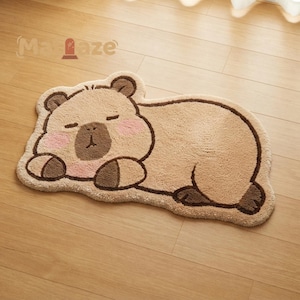 May include: A tan, plush, animal-shaped rug featuring a sleeping capybara with a dark brown nose, dark brown paws, and soft pink cheeks. The rug has a thick, textured pile and a dark brown outline, with the text "Ma-laze" visible above.