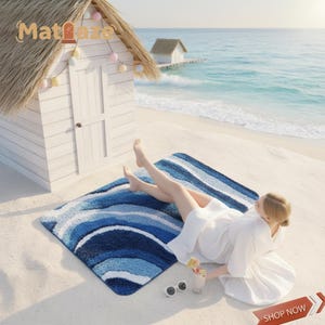 Könnte beinhalten: Eine blau-weiß gestreifte Strandmatte mit einer Person, die sich am Strand entspannt. Die Matte hat ein wellenförmiges Design mit Blautönen. Eine weiße Strandhütte und das Meer sind im Hintergrund. Der Text "Matlaze" ist sichtbar.