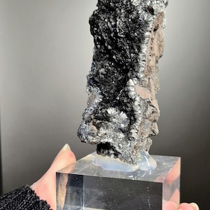 May include: A dark, textured mineral specimen, primarily black with silver-gray metallic luster, mounted on a clear acrylic cube. The mineral has a rough, irregular surface and is held by a hand.