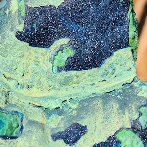 May include: Close-up of a mineral specimen with a dark blue, sparkling center surrounded by light green and blue-green formations. The surface has a textured, rough appearance, with variations in color and depth.