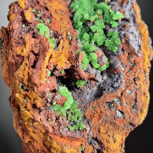 Op de afbeelding: Close-up van een mineraalmonster met een ruw, getextureerd oppervlak. De steen heeft een mix van kleuren, waaronder roestoranje, geel, grijs en vlekken van levendige groene kristalformaties. De algehele uitstraling is natuurlijk en aards.
