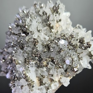 Può includere: Primo piano di un gruppo di cristalli di quarzo con inclusioni di pirite. I cristalli sono chiari e traslucidi, con una lucentezza metallica scintillante dalla pirite. Il colore generale è un mix di trasparente, argento e oro.