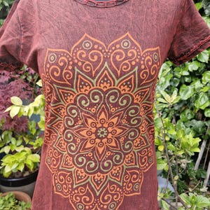 May include: A short-sleeved t-shirt in a dark reddish-brown acid wash finish featuring a large, intricate mandala graphic printed in shades of orange and olive green. The garment has a round neckline with decorative stitching along the collar and sleeves.
