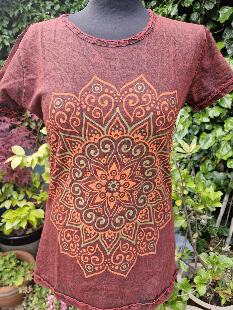 May include: A short-sleeved t-shirt in a dark reddish-brown acid wash finish featuring a large, intricate mandala graphic printed in shades of orange and olive green. The garment has a round neckline with decorative stitching along the collar and sleeves.