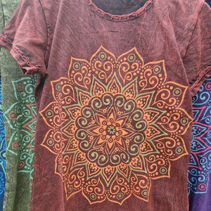 May include: A short-sleeved, dark red t-shirt with a distressed, washed-out texture. The front features a large, intricate mandala graphic printed in shades of orange and olive green. The neckline and hem are finished with visible, decorative stitching.