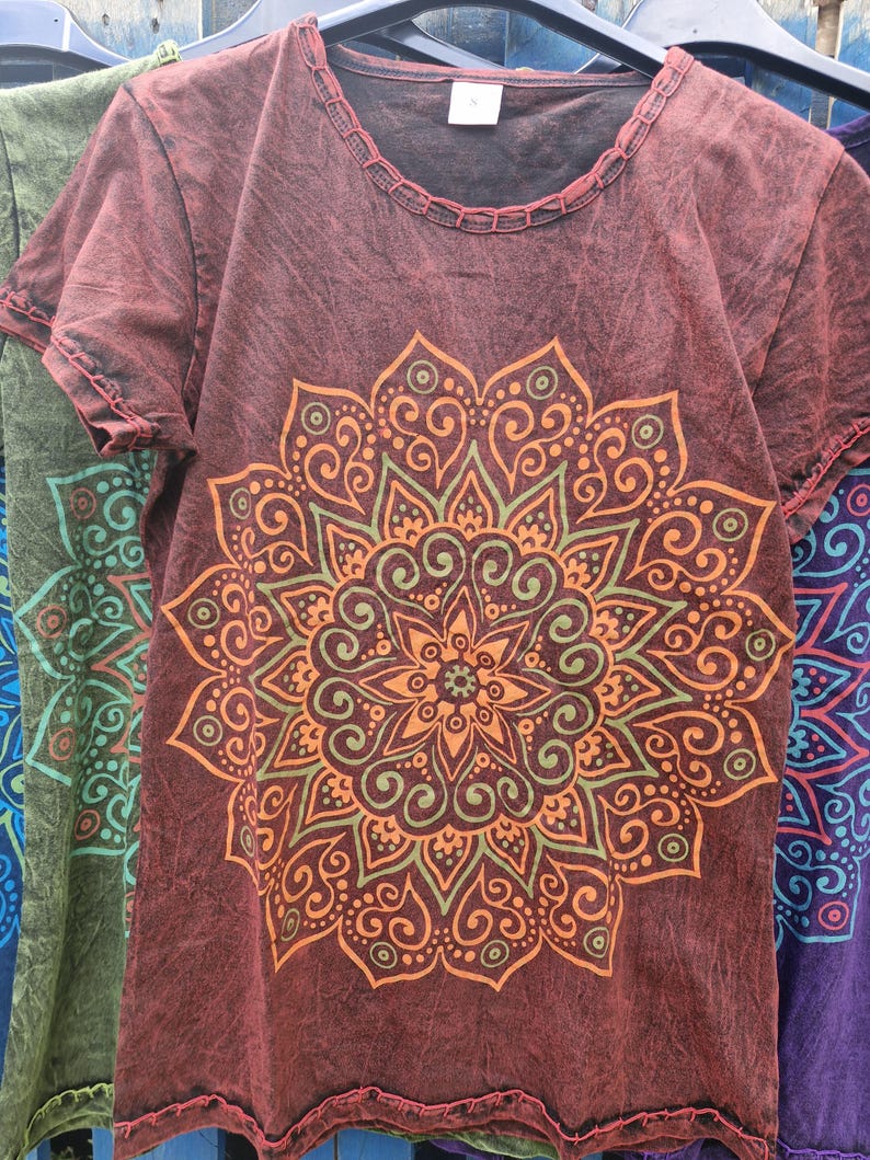 May include: A short-sleeved, dark red t-shirt with a distressed, washed-out texture. The front features a large, intricate mandala graphic printed in shades of orange and olive green. The neckline and hem are finished with visible, decorative stitching.