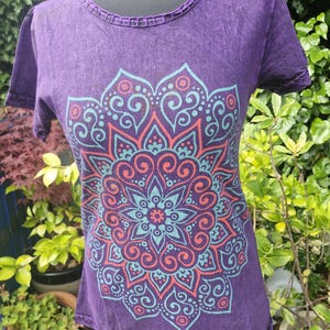 May include: A purple short-sleeved t-shirt featuring a large, intricate mandala graphic printed in shades of teal and coral. The garment has a round neckline with a decorative, open-work stitched trim along the collar and the sleeve edges.