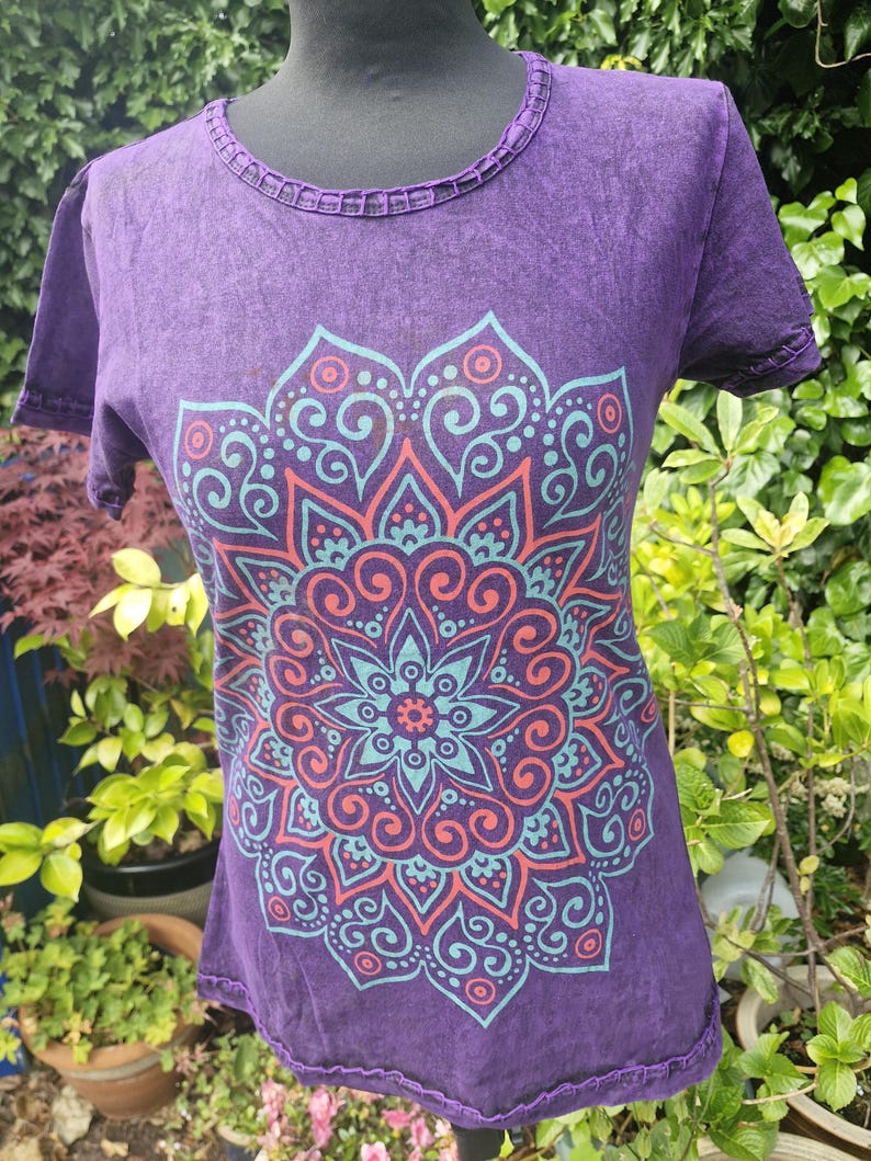May include: A purple short-sleeved t-shirt featuring a large, intricate mandala graphic printed in shades of teal and coral. The garment has a round neckline with a decorative, open-work stitched trim along the collar and the sleeve edges.
