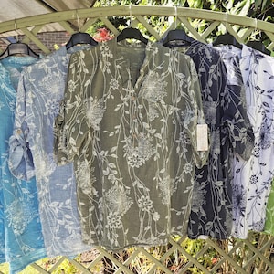 May include: Several floral print shirts in shades of blue, green, and grey are displayed on a rack. Each shirt features a button-down design and rolled-up sleeves. The shirts are made from a lightweight material.
