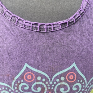 May include: A purple sleeveless top featuring a round neckline with decorative whipstitch detailing. The fabric has a subtle mottled texture and displays a partial graphic print in light blue and coral orange, showcasing a symmetrical floral pattern.