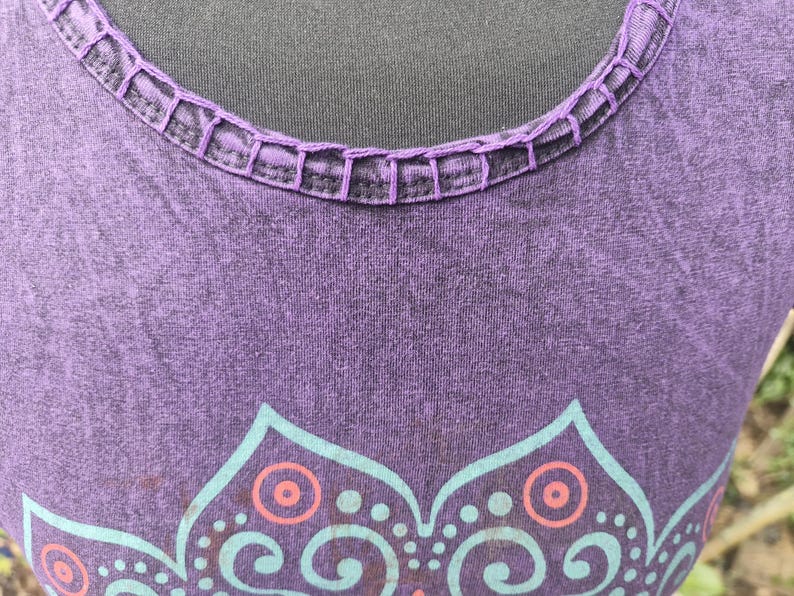 May include: A purple sleeveless top featuring a round neckline with decorative whipstitch detailing. The fabric has a subtle mottled texture and displays a partial graphic print in light blue and coral orange, showcasing a symmetrical floral pattern.
