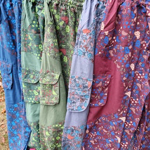 May include: Five pairs of cargo trousers hanging on black hangers, each featuring an elastic waistband and side cargo pockets. The trousers display various colour schemes including blue, green, olive, grey, and maroon, all patterned with mushrooms and flowers.