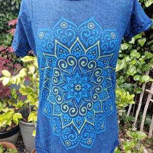 May include: A short-sleeved t-shirt in a deep blue acid-wash finish featuring a large, symmetrical mandala graphic printed in shades of light blue and lime green. The garment has a round neckline with visible stitched detailing along the collar and sleeves.