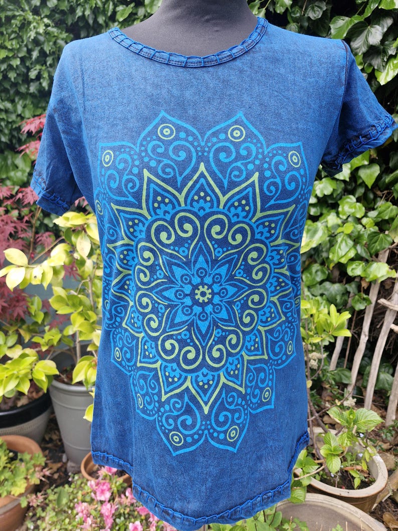 May include: A short-sleeved t-shirt in a deep blue acid-wash finish featuring a large, symmetrical mandala graphic printed in shades of light blue and lime green. The garment has a round neckline with visible stitched detailing along the collar and sleeves.