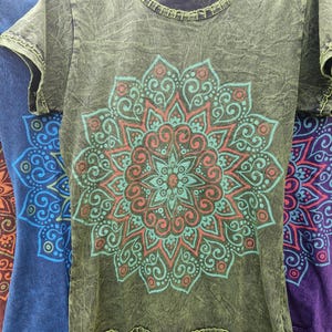May include: An olive green short-sleeve t-shirt with a mineral wash finish and a round neckline. The front features a large, intricate mandala graphic printed in shades of teal and coral, with decorative stitching along the collar and sleeve hems.