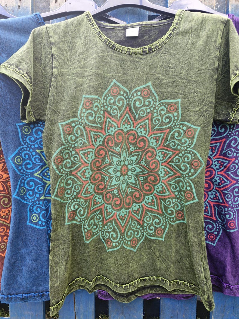 May include: An olive green short-sleeve t-shirt with a mineral wash finish and a round neckline. The front features a large, intricate mandala graphic printed in shades of teal and coral, with decorative stitching along the collar and sleeve hems.