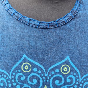 May include: A blue t-shirt with a mottled, washed-out texture and a round neckline featuring decorative stitched detailing. The front of the garment displays a large, symmetrical graphic print in light blue and lime green, featuring intricate floral patterns.