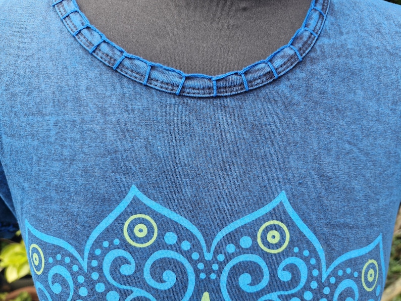 May include: A blue t-shirt with a mottled, washed-out texture and a round neckline featuring decorative stitched detailing. The front of the garment displays a large, symmetrical graphic print in light blue and lime green, featuring intricate floral patterns.