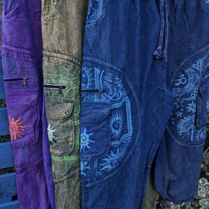 May include: Three pairs of cargo trousers in purple, olive green, and blue, each featuring an elastic waistband with a drawstring, side cargo pockets with zippers, and embroidered sun and celestial patterns on the pockets and leg panels.