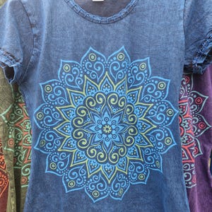 May include: A short-sleeved, acid-wash blue t-shirt featuring a large, symmetrical mandala graphic printed in shades of light blue and lime green. The shirt has a round neckline with visible stitching detail along the collar and sleeve hems.