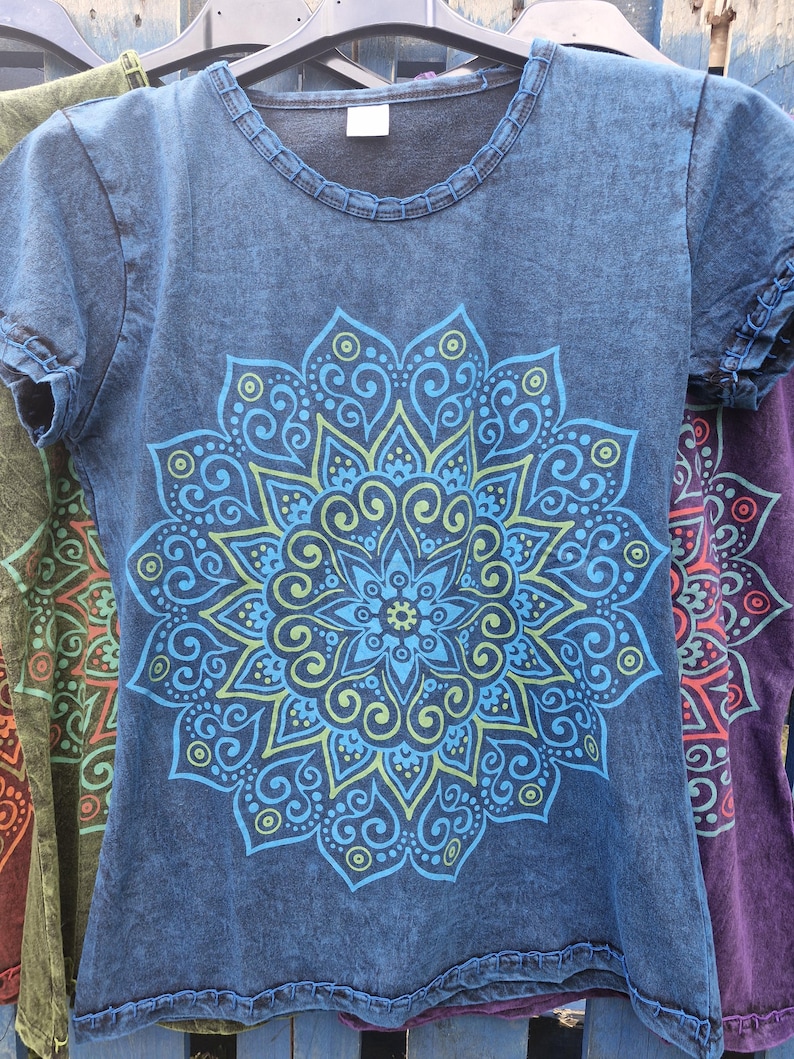 May include: A short-sleeved, acid-wash blue t-shirt featuring a large, symmetrical mandala graphic printed in shades of light blue and lime green. The shirt has a round neckline with visible stitching detail along the collar and sleeve hems.