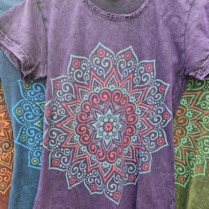 May include: A purple short-sleeved t-shirt with a round neckline and a decorative stitched hem. The front features a large, symmetrical mandala graphic printed in light blue and coral orange, showcasing intricate swirling patterns and shapes.
