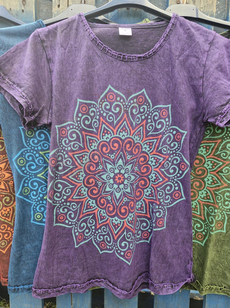 May include: A purple short-sleeved t-shirt with a round neckline and a decorative stitched hem. The front features a large, symmetrical mandala graphic printed in light blue and coral orange, showcasing intricate swirling patterns and shapes.