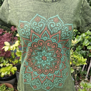 May include: A short-sleeved olive green t-shirt with a crinkled texture and a round neckline. The front features a large, symmetrical mandala graphic printed in shades of teal and coral. The collar and sleeve edges have visible stitching.