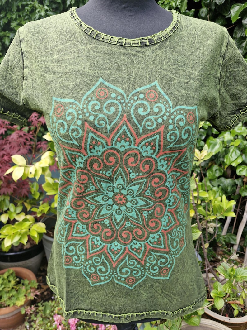 May include: A short-sleeved olive green t-shirt with a crinkled texture and a round neckline. The front features a large, symmetrical mandala graphic printed in shades of teal and coral. The collar and sleeve edges have visible stitching.
