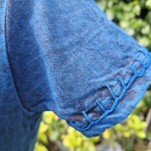 May include: A blue t-shirt sleeve with a heathered texture and a decorative blanket stitch detail along the hem. The stitching is done in a bright blue thread that contrasts with the darker blue fabric of the sleeve, creating a visible edge finish.