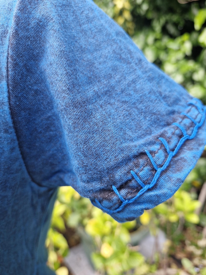 May include: A blue t-shirt sleeve with a heathered texture and a decorative blanket stitch detail along the hem. The stitching is done in a bright blue thread that contrasts with the darker blue fabric of the sleeve, creating a visible edge finish.