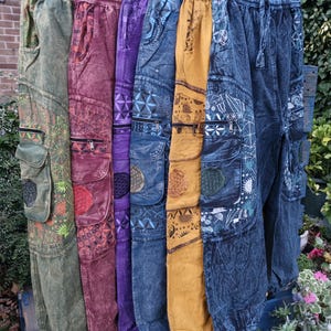 May include: A collection of six pairs of cargo trousers in olive green, maroon, purple, blue, mustard yellow, and dark blue. Each pair features an elastic waistband, side cargo pockets with zippers, and intricate geometric and celestial patterns.