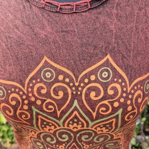 May include: A sleeveless top in a dark maroon, acid-wash fabric featuring a round neckline with decorative stitching. The front displays a large, symmetrical mandala-style graphic print in shades of orange and muted green with intricate patterns.