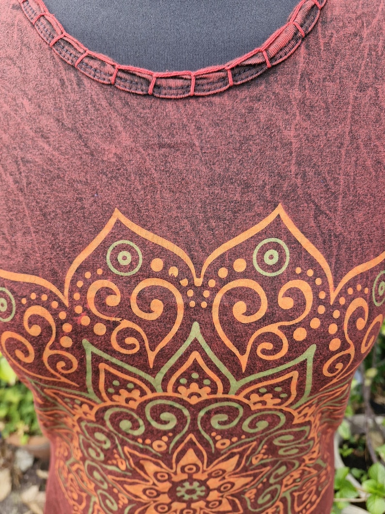 May include: A sleeveless top in a dark maroon, acid-wash fabric featuring a round neckline with decorative stitching. The front displays a large, symmetrical mandala-style graphic print in shades of orange and muted green with intricate patterns.