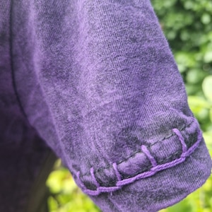 May include: A short-sleeved purple shirt with a heathered texture and a visible decorative blanket stitch detail in a matching purple thread along the edge of the sleeve hem. The fabric has a mottled appearance with darker purple tones.