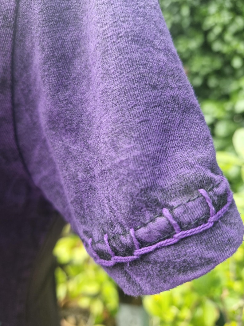 May include: A short-sleeved purple shirt with a heathered texture and a visible decorative blanket stitch detail in a matching purple thread along the edge of the sleeve hem. The fabric has a mottled appearance with darker purple tones.