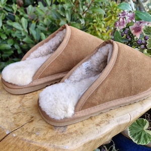May include: A pair of brown suede slippers with a cream-coloured, fluffy interior. The slippers are open-backed and rest on a wooden surface. These comfortable slippers are designed for indoor wear and are ideal for relaxing at home.
