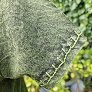 May include: A short-sleeved shirt in a mottled olive green and black acid-wash fabric. The sleeve hem features a decorative blanket stitch detail in a bright lime green thread, creating a distinct contrast against the darker, textured garment fabric.