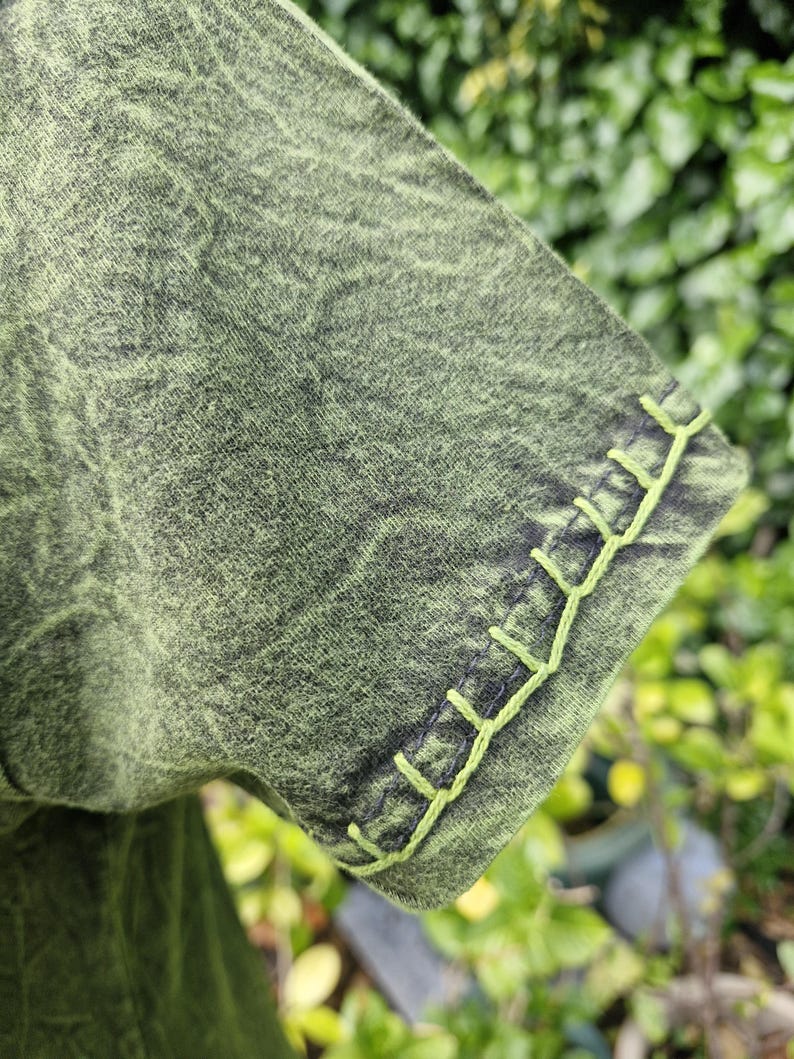 May include: A short-sleeved shirt in a mottled olive green and black acid-wash fabric. The sleeve hem features a decorative blanket stitch detail in a bright lime green thread, creating a distinct contrast against the darker, textured garment fabric.