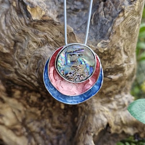 May include: A pendant necklace with an iridescent abalone shell set in a silver frame. The pendant is encircled by red, pink, and blue enamel rings. The necklace hangs from a thin, silver chain. The background is a blurred view of wood.