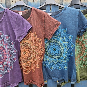 May include: Four short-sleeve t-shirts in purple, brown, blue, and olive green, each featuring a large, intricate, multi-layered mandala graphic print on the front. The fabric has a mottled, acid-wash texture with raw-edge detailing at the neck.