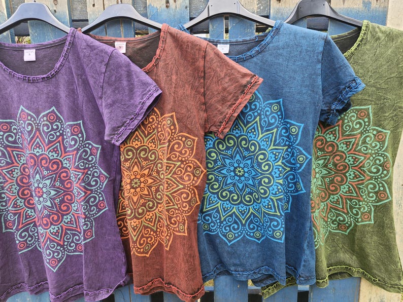 May include: Four short-sleeve t-shirts in purple, brown, blue, and olive green, each featuring a large, intricate, multi-layered mandala graphic print on the front. The fabric has a mottled, acid-wash texture with raw-edge detailing at the neck.