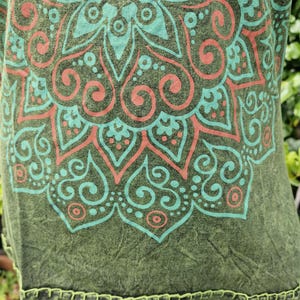 May include: An olive green shirt featuring a large, intricate mandala pattern printed in light teal and coral orange. The fabric has a mottled, textured appearance, and the bottom hem is finished with visible, decorative light green stitching.