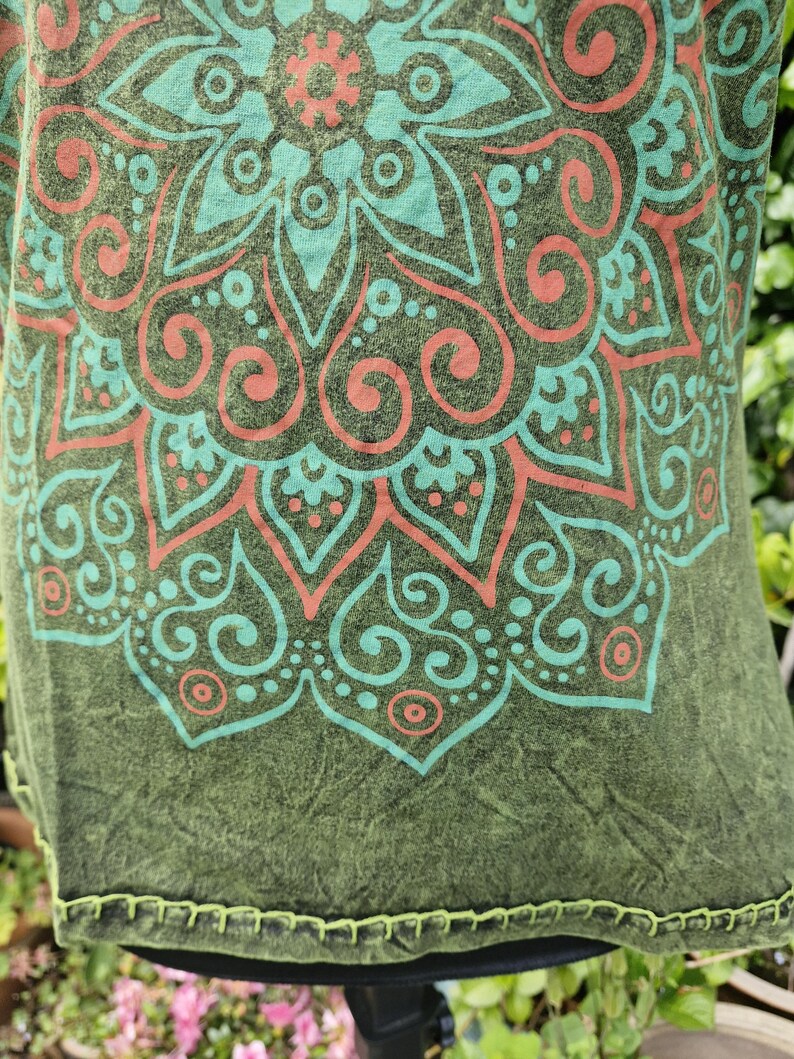 May include: An olive green shirt featuring a large, intricate mandala pattern printed in light teal and coral orange. The fabric has a mottled, textured appearance, and the bottom hem is finished with visible, decorative light green stitching.
