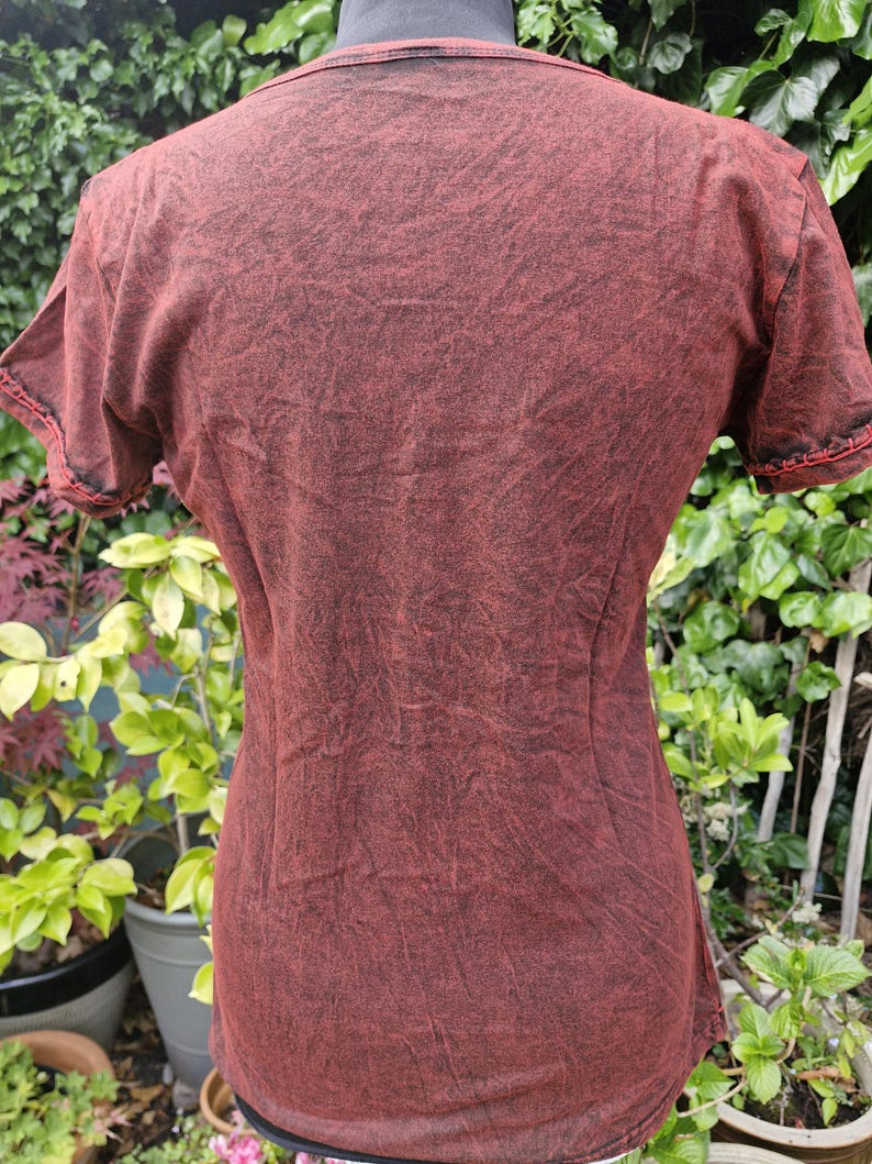 May include: A short-sleeved t-shirt featuring a dark red and black acid-wash pattern. The garment has a round neckline and visible stitching along the sleeve hems. The fabric displays a mottled, textured appearance throughout the entire surface.