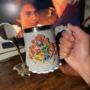May include: A gray, textured mug featuring the Hogwarts crest with a silver rim. The crest is in full color with the house symbols. The handle is a sculpted, gray design. The mug is held in a hand.