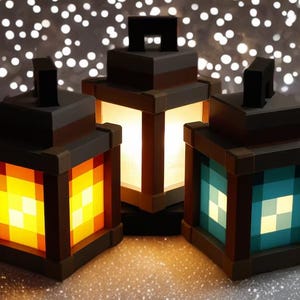 May include: Three illuminated lanterns with a pixelated design. The lanterns have a dark brown frame with orange, white, and teal-colored light panels. Each lantern has a black handle on top. The background is a bokeh effect.