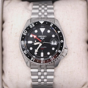 May include: A stainless steel Seiko 5 automatic GMT watch with a black dial and a silver band. The watch features a rotating bezel with white numerals and a red GMT hand. The watch is displayed on a white pillow.