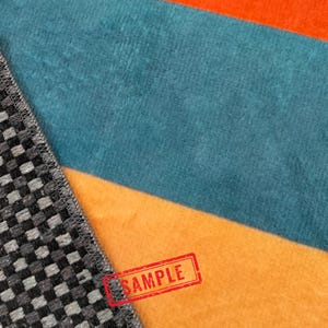 May include: Close-up of a colorful textile with a geometric pattern and a "SAMPLE" stamp. The fabric features stripes of orange, teal, and a dark gray and black checkered pattern.